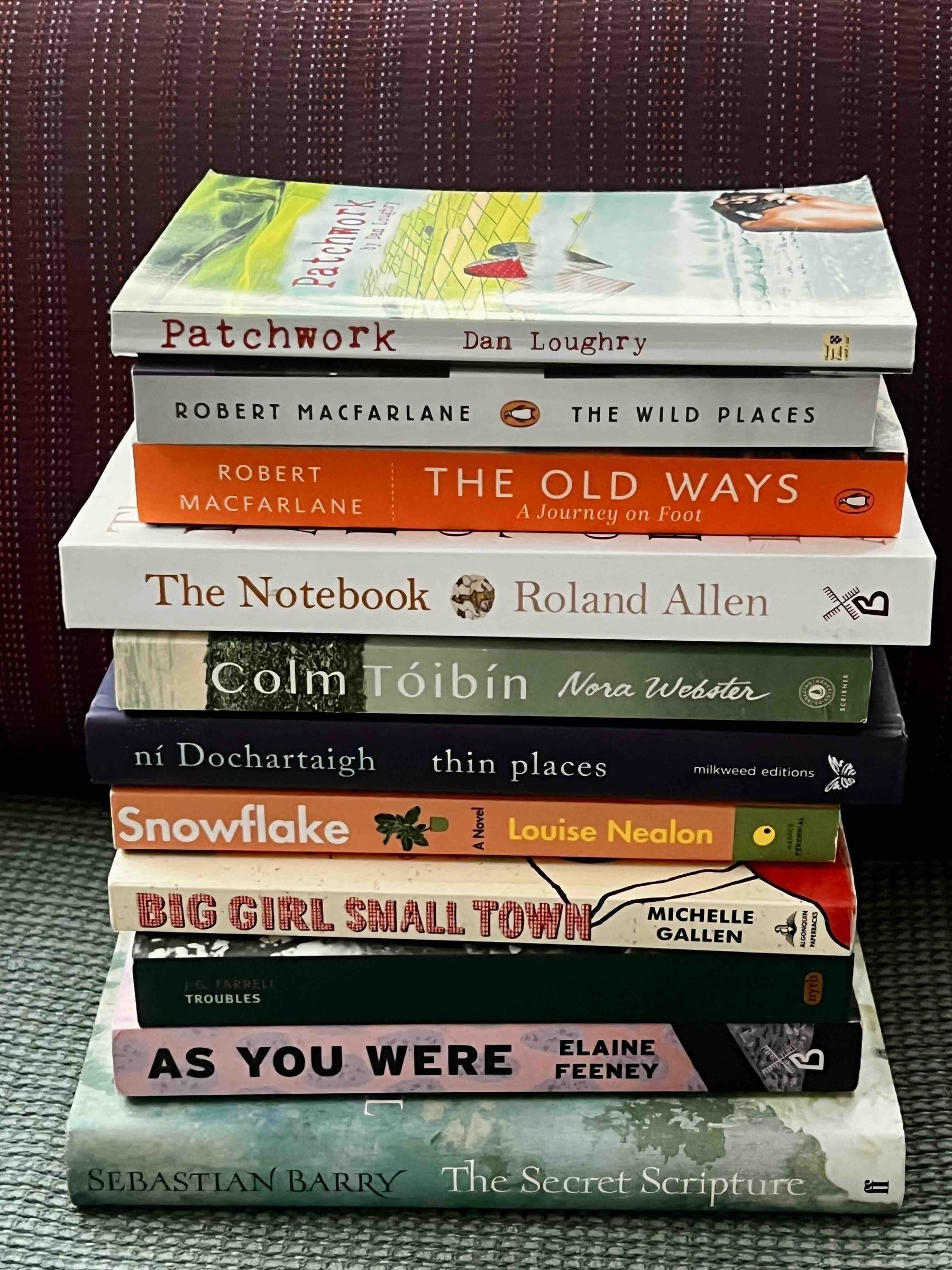 A stack of nine books with various authors and titles, including "Patchwork" by Dan Loughry and "The Secret Scripture" by Sebastian Barry, organized on a purple upholstered surface.